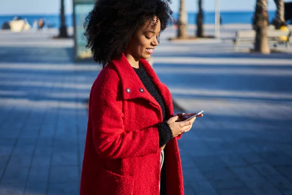 携帯電話を見ている間に舗装されて立って暖かい服の若いアフリカ系アメリカ人女性の側のビューと喜んで笑顔 — ストック写真
