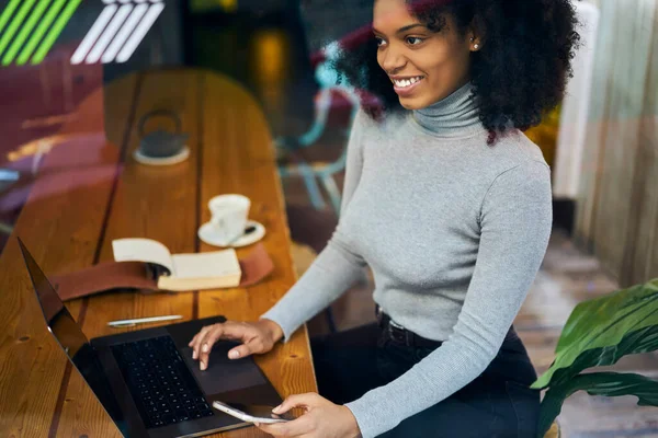 明るいアフリカ系アメリカ人女性のガラスを通して高い角度カジュアルウェアノートパソコンとブラウジングスマートフォンと木製のテーブルに座って — ストック写真
