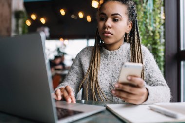 Akıllı telefon aygıtı ile Afrika kökenli Amerikalı kız öğrenci sınav hazırlıkları için öğrenim sırasında bilgilendirici web sitesini araştırıyor. Laptop 'ta ders kitabı okuma eğitimi alan yetenekli bir kadın.