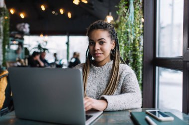 Koyu tenli kadın milenyum portresi kafeterya masasında modern dizüstü bilgisayarla oturuyor ve kameraya bakıyor, yetenekli serbest grafik tasarımcısı netbook kullanarak uzaktan iş yapıyor.