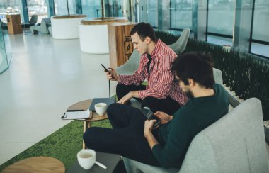 Yukarıdan bakıldığında, rahat koltuklarda otururken ve modern çalışma alanlarında dinlenirken akıllı telefonlar kullanan ve kahvenin tadını çıkaran konsantre genç erkek iş arkadaşları görülüyor.