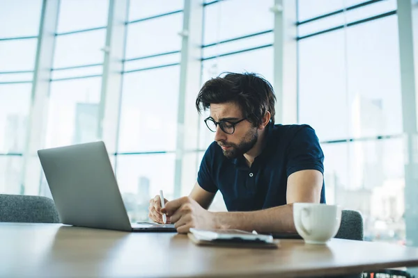 严肃的年轻男子 留着深色胡子 穿着休闲装 坐在桌旁 拿着笔记本电脑和咖啡 在当代工作场所制定计划 — 图库照片