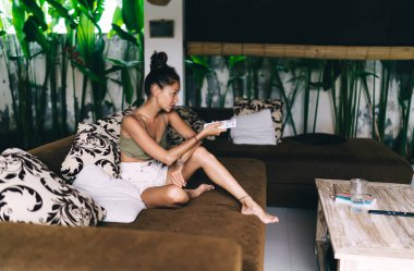 Yandan bakıldığında, rahat bir kanepede, duvar dikişçilerine karşı dinlenen ve uzaktan kumandayla kanal değiştiren gündelik kıyafetli bir etnik kadın.