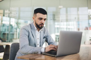 Modern netbook klavyesinde yazarken çağdaş ofiste cam duvarlara karşı çalışan şık takım elbiseli, ciddi, genç ve sakallı bir girişimci.