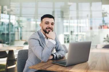 Neşeli, saygıdeğer genç girişimci resmi takım elbiseli netbook ile masada oturmuş modern iş merkezinde cam duvara bakarken çenesine dokunuyor.