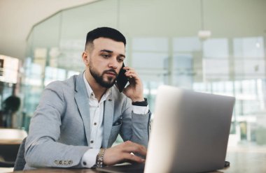 Düşünceli genç erkek yönetici gri takım elbise giyip internet sayfalarına göz atıyor ve modern iş merkezinde cam duvarlarla çalışırken cep telefonuyla konuşuyor.