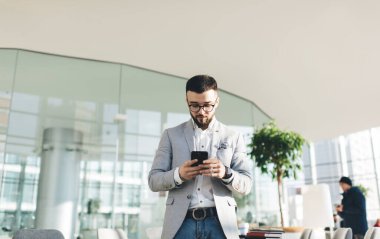 Modern bina terasında güneşli bir günde cam duvara yaslanırken resmi olarak erkek ofis yöneticisi cep telefonuyla mesajlaşır.