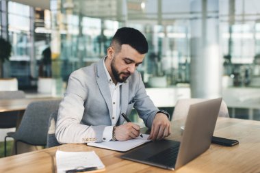 Zarif takım elbiseli genç ve yakışıklı bir işadamı geniş bir ofiste dizüstü bilgisayarla masada otururken deftere bir şeyler yazıyor.