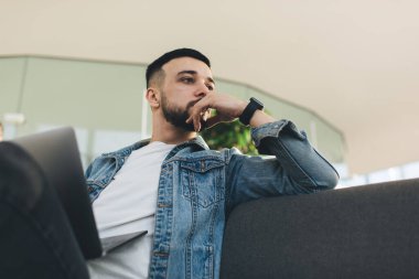 Kanepede dizüstü bilgisayarla oturan sakallı, ciddi bir genç adam. Parlak salonda çalışırken iş meselelerini düşünüyor.