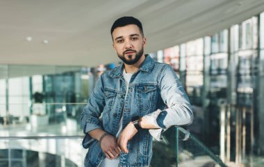 Sakallı, modaya uygun ceketli, koridorda dikilip cam korkuluklara yaslanan ve kameraya bakan adam.