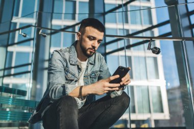 Modern binadaki cam merdivenlerde oturan ve tatilde akıllı telefonları karıştıran sıradan kıyafetli ciddi sakallı bir adamın alçak açısı.