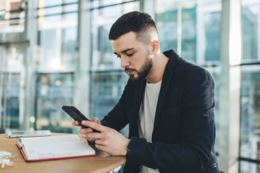 Kendine güveni tam sakallı bir adam ahşap masada elinde defterle oturuyor ve modern kafede çalışırken akıllı telefon sörfü yapıyor.