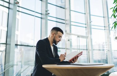 Resmi kıyafetli, düşünceli, sakallı bir erkek, ahşap masada dizüstü bilgisayarla oturuyor ve kafede kitap okuyor.