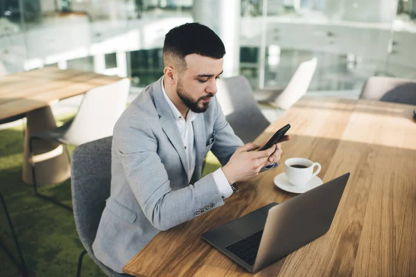 在当代玻璃工作场所 穿着灰色高级西装的男性经理一边坐在桌旁一边浏览智能手机 一边与上网本和一杯热的美式咖啡坐在一起 — 图库照片