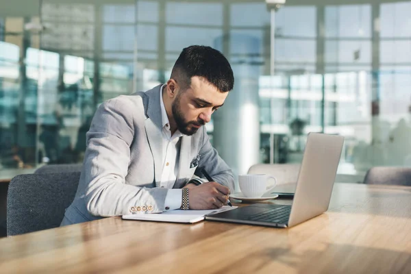 在现代工作空间工作时 专注的大胡子商人穿着正式服装 带着笔记本电脑在桌旁工作 并在笔记本上写笔记 — 图库照片