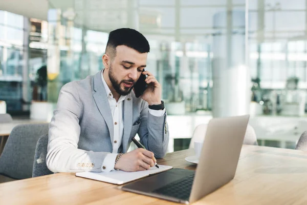 在现代办公室里 年轻英俊的商人一边坐在办公桌前 一边用手机聊天 一边用笔记本写字 — 图库照片