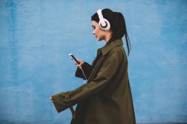 Paltolu, siyah saçlı, akıllı telefonlu, kulaklıklı, mavi arka planda müzik dinleyen şık bir kadın.