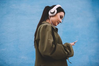 Mavi arka planda kulaklıkla müzik dinlerken ceketli, gülümseyen ve cep telefonlu neşeli genç bayanın yan görüntüsü