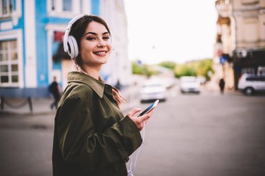 Yandan gülümseyen kahverengi saçlı kadın kulaklıkla müzik dinlerken akıllı telefon kullanıyor ve kameraya bakıyor.