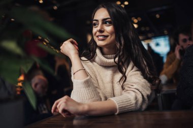 Kahvede sıcak bej kazaklı, mutlu, genç, siyah saçlı bayan dişleri sırıtırken ve yüzünü çevirirken masada oturuyor.