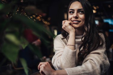 Rahat örülmüş süveter giyen, elleri çenesinde gülümseyen, rahat bir kafede hayal kuran ve gözlerini kaçıran mutlu genç bayan.