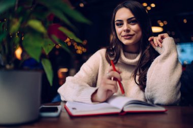 Konsantre olmuş, rahat bir süveter giymiş, elinde not defteriyle masada oturan ve elinde kalem ışığı olan mutlu bir kadın.