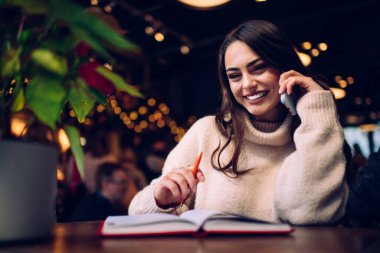 Gülümseyen esmer kadın, turuncu kalemle ve planlamacıyla kahkahalarla arka tarafta dinlenirken cep telefonuyla gülüyor ve konuşuyor.