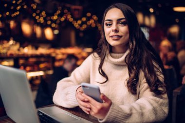 Karizmatik, uzun saçlı, günlük kıyafetlerle gezen akıllı telefonlu bir kadın. Modern kafede bilgisayarla çalışırken kameraya bakıyor.