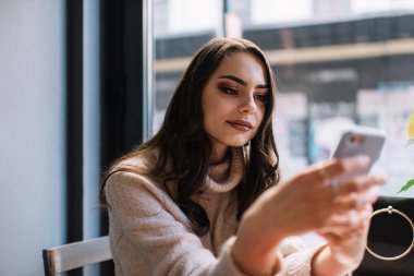 Modern bir dairede boş vakit geçirirken uzun saçlı, ciddi bir genç kadın akıllı telefondan video izliyor.