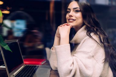 Kafede uzaktan çalışırken ve camdan dışarı bakarken gündelik kıyafetlerle gezen çekici kahverengi saçlı kadın.