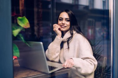 İyimser modaya uygun esmer, rahat bir kafenin içinde oturmuş dizüstü bilgisayarla uzaktan çalışırken iş projesini düşünüyor.