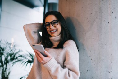 Kafede beton duvara yaslanırken akıllı telefon kullanan kazaklı ve modern gözlüklü neşeli bir kadın.