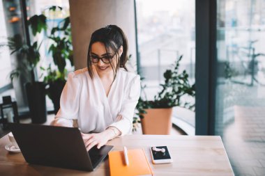 Kafede vakit geçirirken tahta masada oturup dizüstü bilgisayarda sörf yapan, günlük giysiler ve gözlüklerle serbest çalışan neşeli kadın.