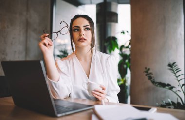 Beyaz bluzlu, ciddi bir bayan patron. Düşünceli bir şekilde kafasını çeviriyor ve elinde bardak ve bardak bardak ile bir yandan da iş yaparken bir yandan da içki bardağı tutuyor.