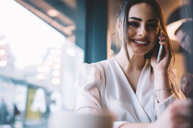 Modern ışıklı kafede büyük pencerenin yanında oturan ve cep telefonuyla konuşan şık beyaz elbiseli genç bayan.