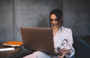 Modayı takip eden beyaz bluzlu, gözlüklü, dizüstü bilgisayarlı, internette gezen ve gülümseyen genç bir kadın.