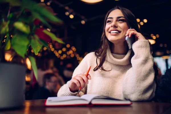 Akıllı telefon tutarken ve telefonda gülerken açık not defteri ve kalemle masada oturan neşeli kadın.