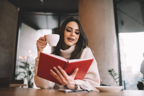 暖かい服の正の女性の低角度コーヒーを飲みながら本を読みながら カフェで木製のテーブルに座って — ストック写真