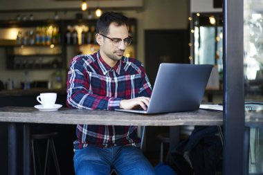 Konsantre sakallı erkek, günlük kıyafet ve gözlüklü. Uzaktan kumandalı bir projede çalışıyor. Rahat bir kafede vakit geçiriyor.