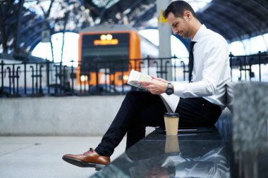 Tüm vücudun yan görünüşü resmi giysiler içinde düşünceli bir erkek istasyonda bankta oturuyor ve teneffüste gazete okuyor.