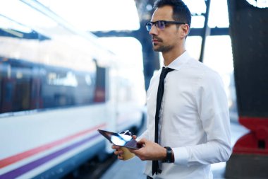 Trenin yanında elinde tablet ve bir fincan kahveyle demiryolu platformunda duran resmi kıyafetler ve gözlüklerle ciddi bir erkek ofis çalışanının yan görüntüsü.