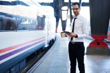 Resmi giysiler ve gözlüklerle tren istasyonunda kahve ve internet tableti taşırken tren bekleyen zarif yetişkin girişimci.