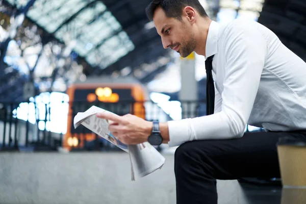 Kendine güvenen, resmi takım elbiseli, kenarda oturmuş tren beklerken gazete okumaktan zevk alan girişimcinin yan görüntüsü.