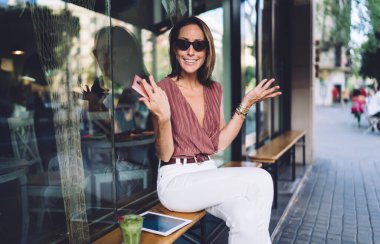 Moda kıyafetli, güneş gözlüklü, gülümseyen yetişkin bir kadın modern kafenin dışındaki cam duvarın yanında oturmuş buzlu içecekle ve omuz silkerek rahatlarken tablet ve akıllı telefonuyla oturuyor.