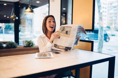 Beyaz gömlekli, gazete okuyan, akıllı telefon ve tabletle masa başında oturan ve modern kafede ağzı cam olan kameraya bakarken hayran olan hoş bir kadın.