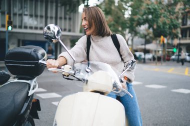 Sırt çantalı neşeli ve mutlu yetişkin bir kadın. Gülümsüyor ve yüzünü çeviriyor. Şehir caddesinde scooter 'da otururken.