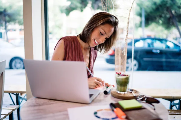 ノートパッドにメモを書いている女性の笑顔カフェでデトックスのガラスと朝食を持っている間 ラップトップ上で作業中に — ストック写真