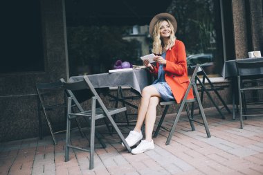 Güzel moda blogcusu şık giyinmiş dijital tableti elinde tutuyor ve haftasonu kaldırım kenarında eğlenirken gülümsüyor. Dokunma yastığı olan mutlu beyaz kız.
