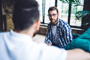 Modern gözlüklü ciddi sakallı bir adam iş arkadaşıyla konuşuyor. Gün ışığında rahat bir iş yerinde proje üzerinde birlikte çalışıyorlar.