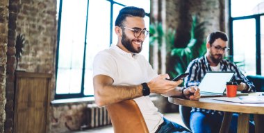 Uzaktan kumandalı meslektaşı ile bilgisayar programlama için yaptığı işbirliği sırasında akıllı telefon sohbetinin keyfini çıkaran mutlu Türk bilişim uzmanı, göz giysisi giyen neşeli hippi adam komik metin yayınları okuyor
